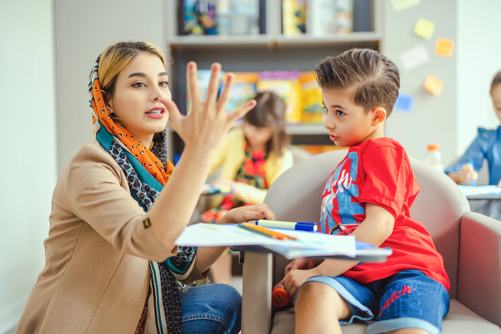 A teacher counts on her fingers with two young students at their classroom table.